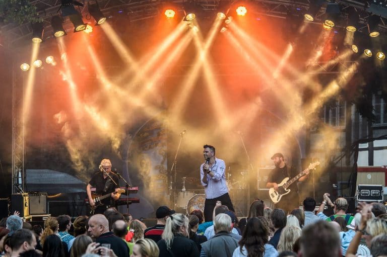 Mitglieder der Coverband Abenteuerland performance auf einer Bühne bei einem Open-Air-Konzert, umgeben von Publikum, mit bunten Scheinwerfern und Rauch für eine unvergessliche Musikveranstaltung in Deutschland.