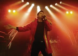 Abenteuerland Coverband bei einem Live-Auftritt, leidenschaftlicher Sänger in rotem Outfit vor farbenfroher Bühnenbeleuchtung, perfekte Stimmung für unvergessliche Abendveranstaltungen, Hochzeitsfeiern und Festivals.