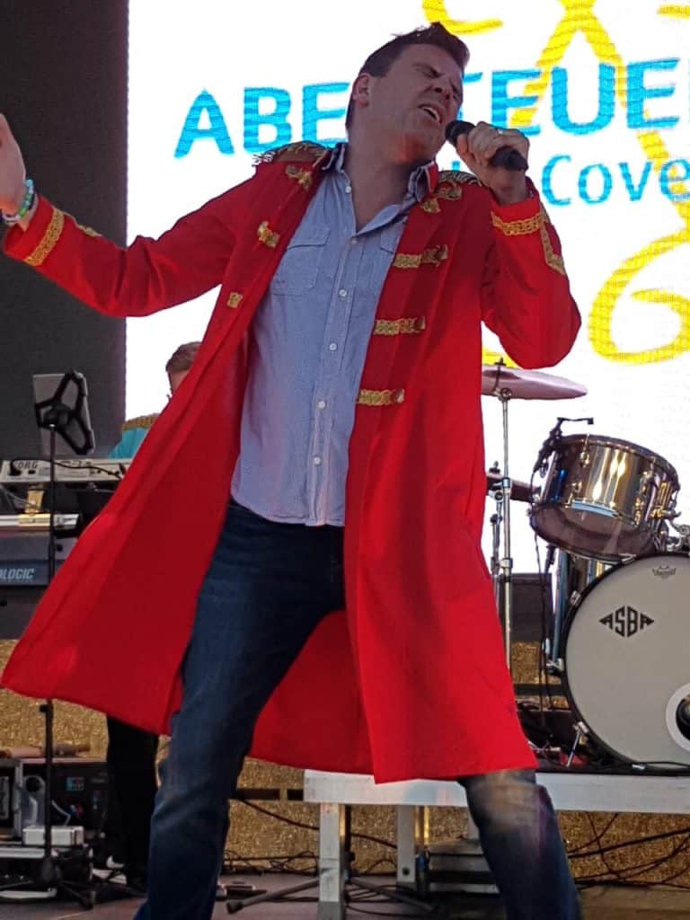 Abenteuerland Coverband Live-Performance, Sänger im roten Ordenkostüm, energiegeladene Show mit Musik und Trommel auf Bühne.