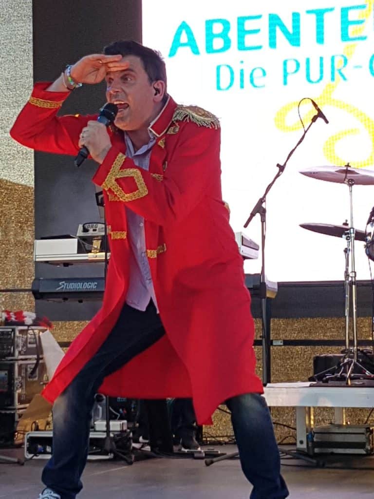 Abenteuerland Coverband Performance beim Event, Musiker in rotem Jackett, singend und mit Hand am Kopf, Bühnenausstattung, große LED-Leinwand im Hintergrund, Entertainer auf der Bühne, Live-Musik Show, Spaß und Energie, professionelle Liveband für Events und Partys.